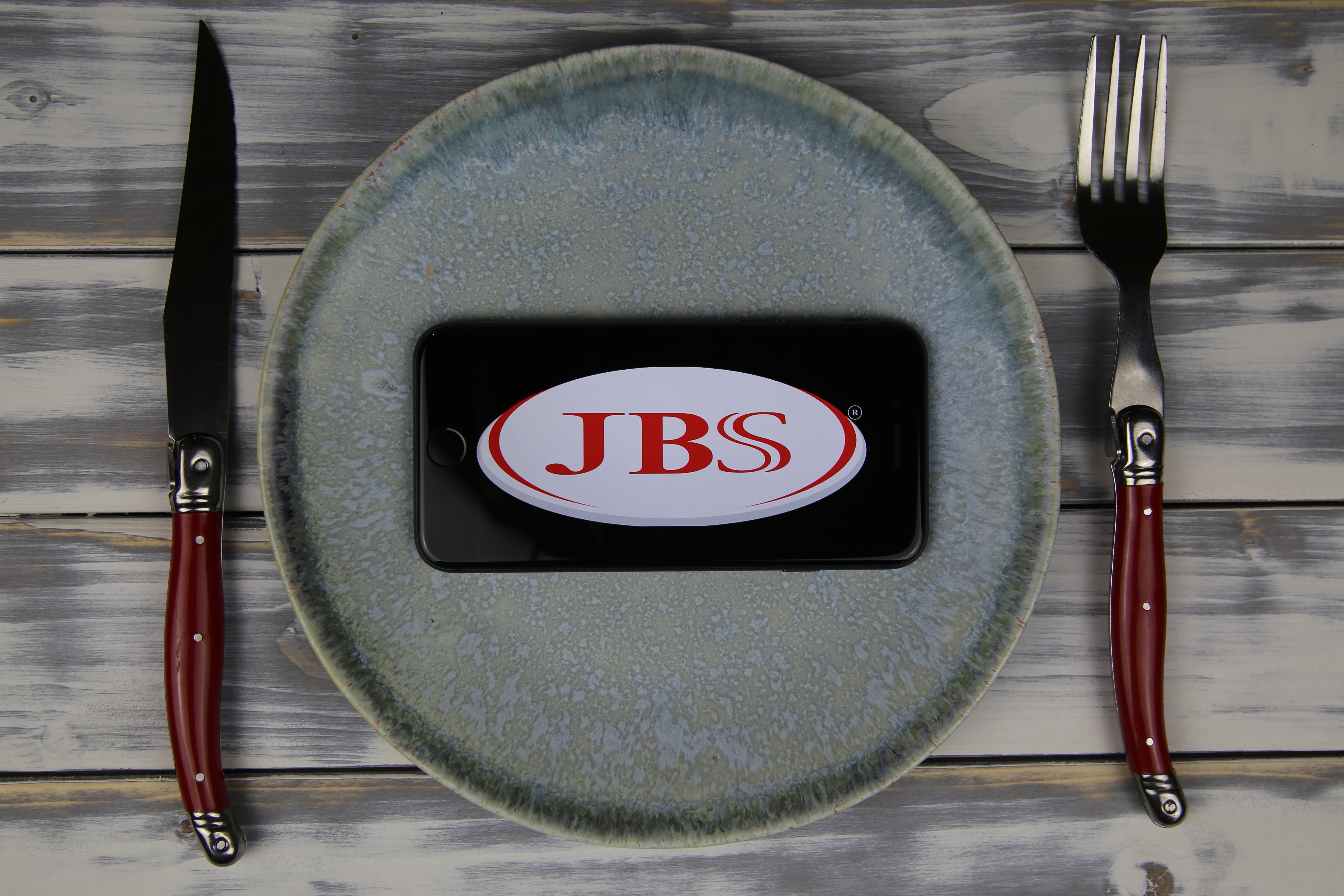 Viersen, Germany - May 9. 2020: Closeup of mobile phone on plate and wood table, cutlery with logo lettering of Jose batista sobrinho JBS food company (focus on center)